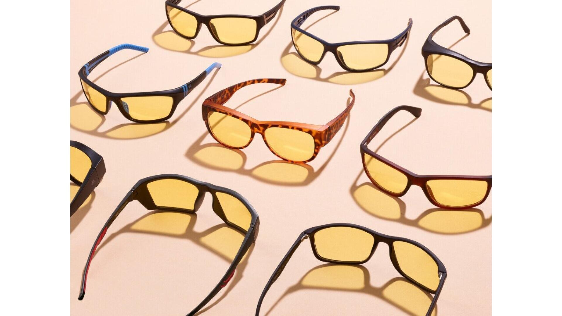 Several pairs of sunglasses with yellow-tinted lenses and dark or tortoiseshell frames are arranged on a peach-colored surface, casting soft shadows.