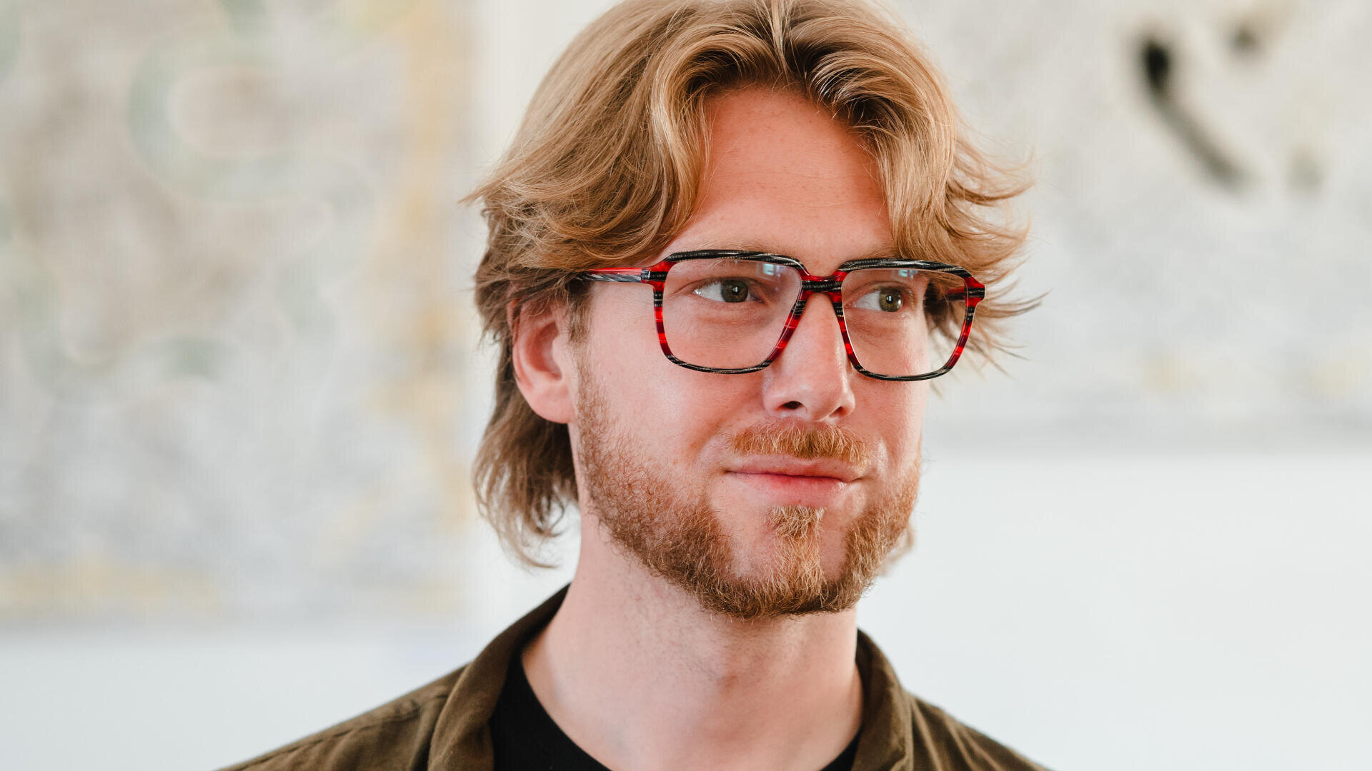 Ein junger Mann mit welligem blondem Haar, kurzem Bart und einer roten Schildpattbrille blickt leicht zur Seite. Er trägt ein dunkles Hemd und eine Jacke, im unscharfen Hintergrund ist ein abstraktes Kunstwerk zu sehen.