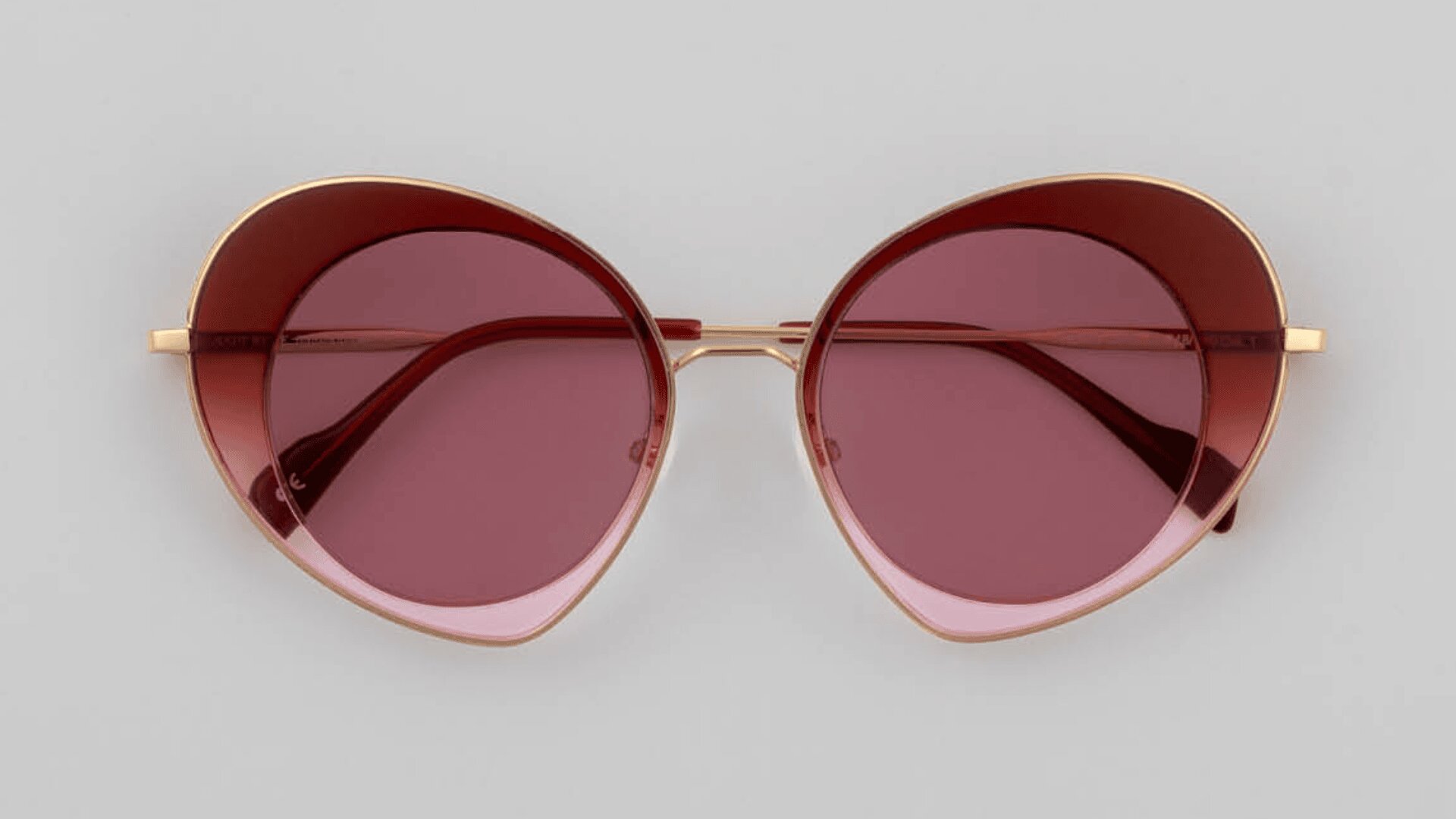 Rot getönte Cat-Eye-Sonnenbrille mit goldenem Rahmen auf hellgrauem Hintergrund.