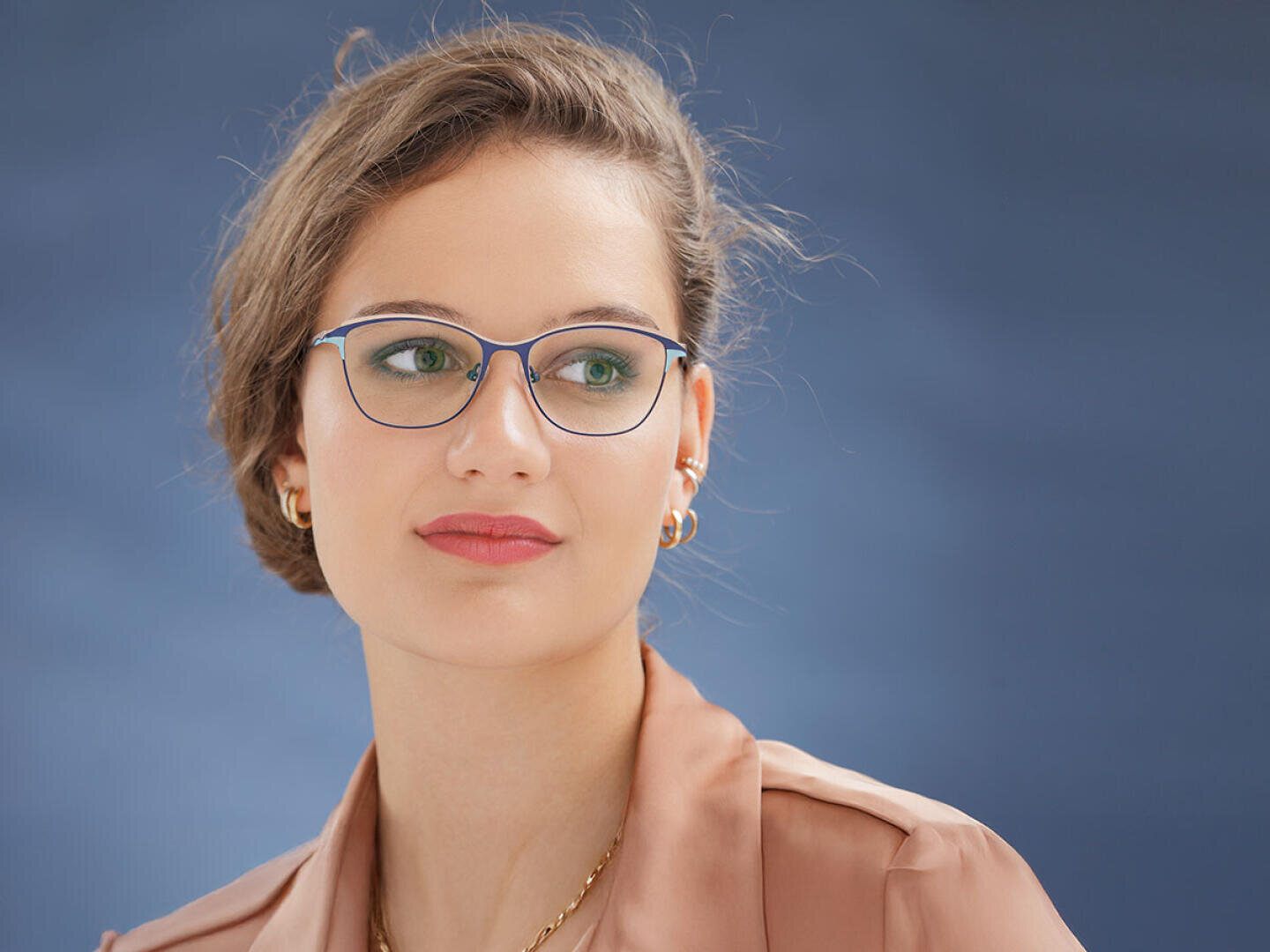 A woman with fair skin, brown hair styled to one side, glasses, gold earrings and a light brown blouse looks thoughtfully to the left against a blue background.