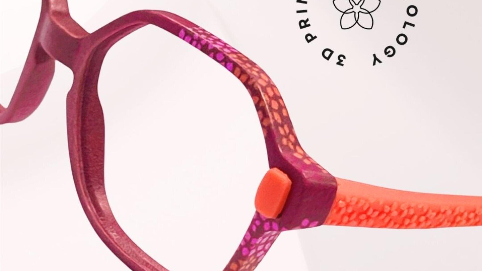 Close-up of colorful eyeglass frames with a mix of purple, orange, and pink hues and a textured pattern. In the background, there is a round black logo featuring the words “3D PRINTED” and a floral design.