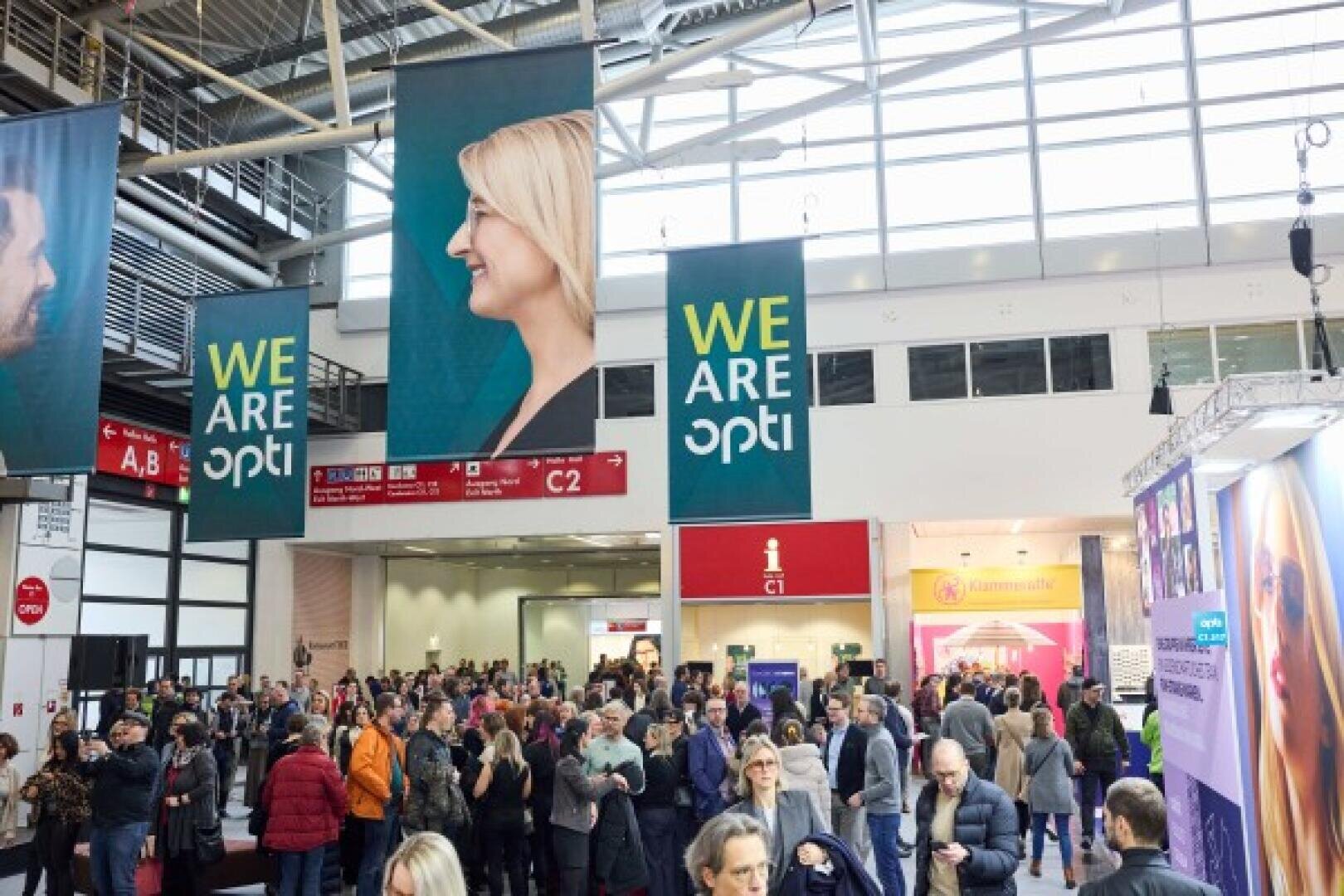 Eine große Menschenmenge versammelt sich in einem Kongresszentrum unter Bannern mit der Aufschrift „WIR SIND opti“, im Hintergrund sind Ausstellungsstände und Beschilderungen zu sehen.