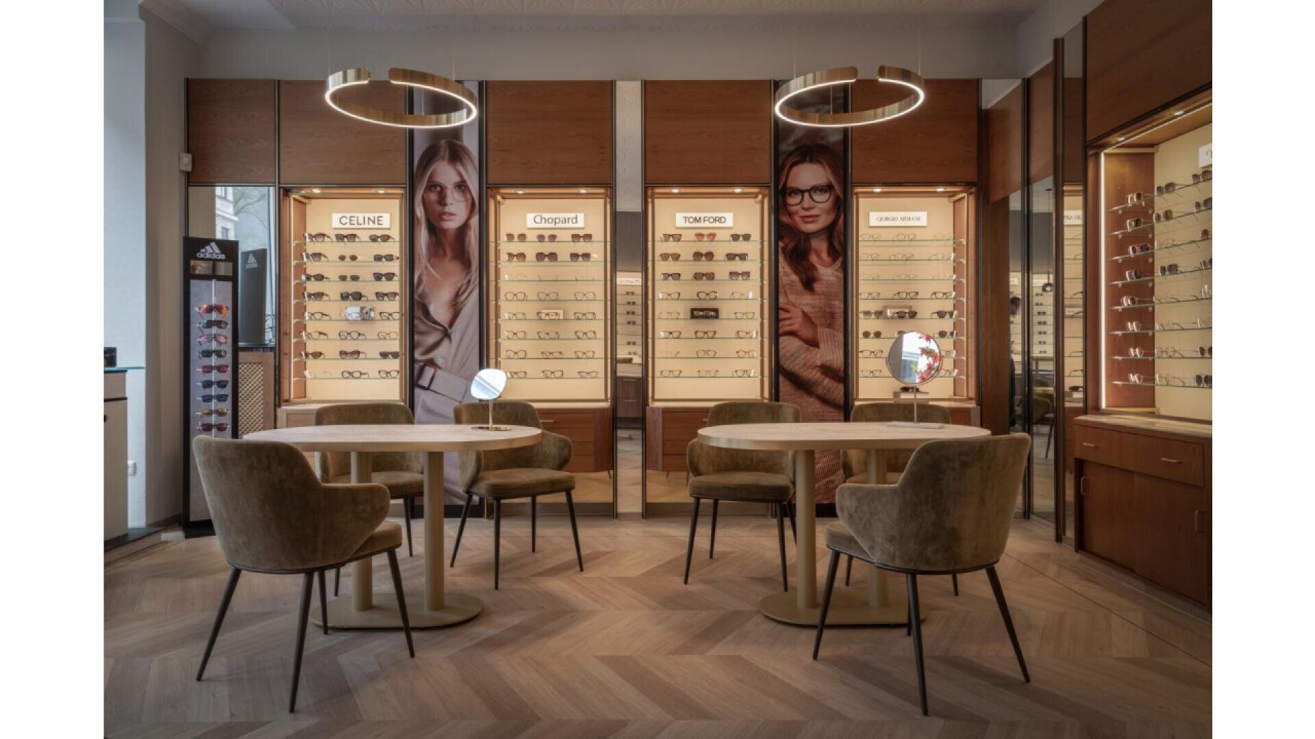 A stylish eyewear store interior with wooden floors, two round tables with chairs, display shelves of glasses, and large posters of women wearing eyeglasses on the back wall. Two circular ceiling lights hang above.