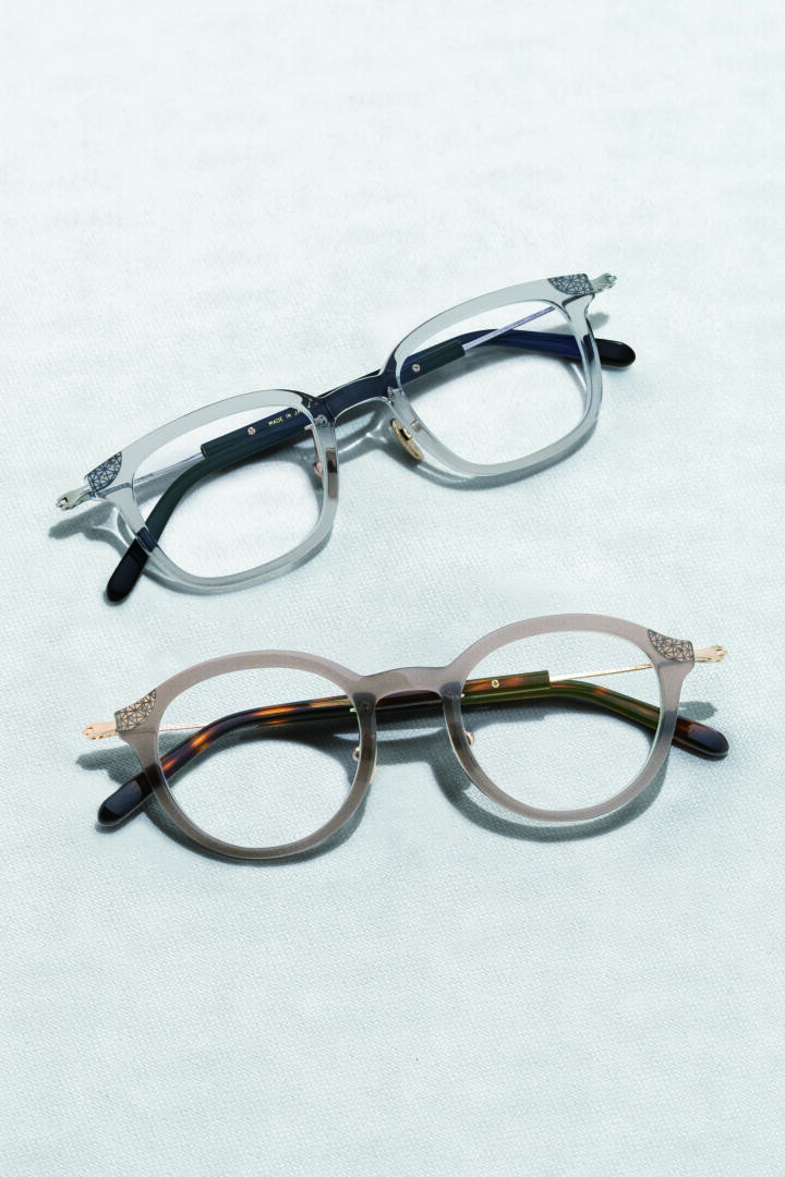 Two pairs of eyeglasses with clear and tortoiseshell frames are placed on a light textured surface, one pair above the other.