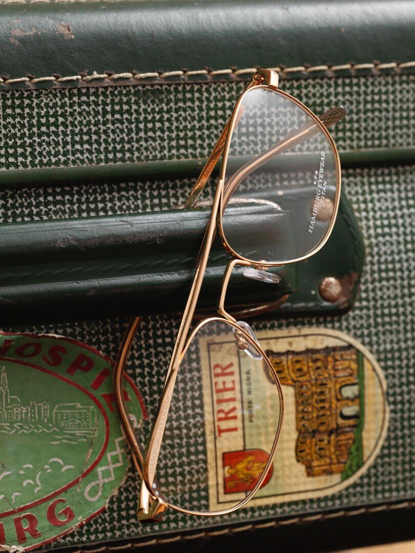 Eine goldfarbene Brille liegt auf einem grünen Koffer, der mit Reiseaufklebern verziert ist, unter anderem mit den Aufschriften Trier und Hospiz.