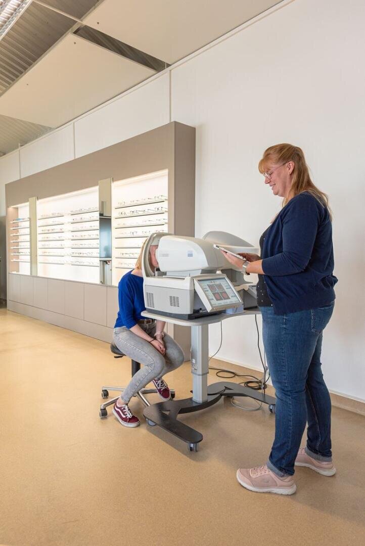 A woman operates an eye examination machine while a seated girl looks into the device in an optician's store, while spectacle frames hang on the wall in the background.