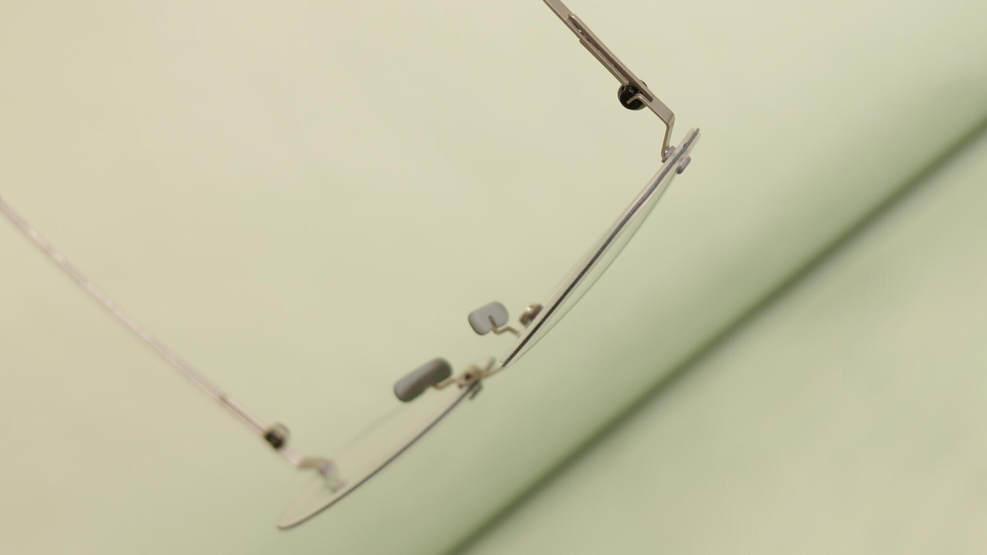 Nahaufnahme des Rands und der Nasenpads einer Brille, die schräg vor einem weichen, hellgrünen Hintergrund gehalten wird.
