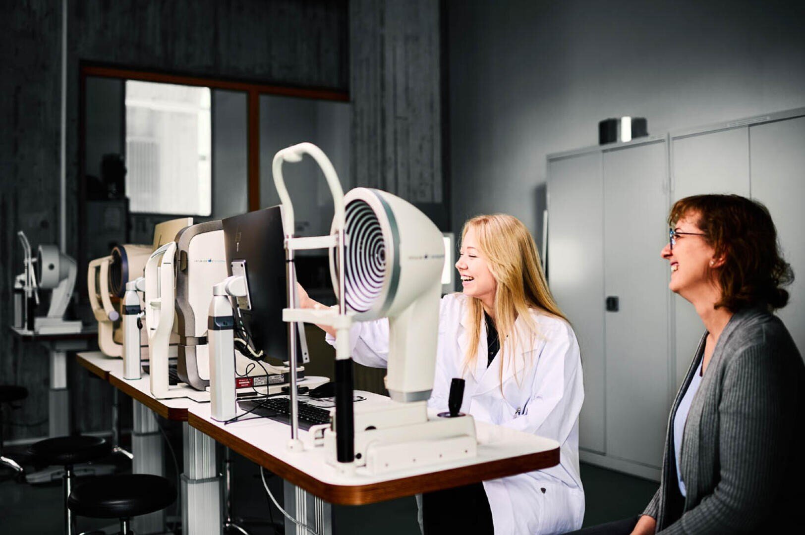 Two women sit at a row of eye examination machines; one wears a white lab coat and operates a machine while both smile, in a bright and modern clinical environment.