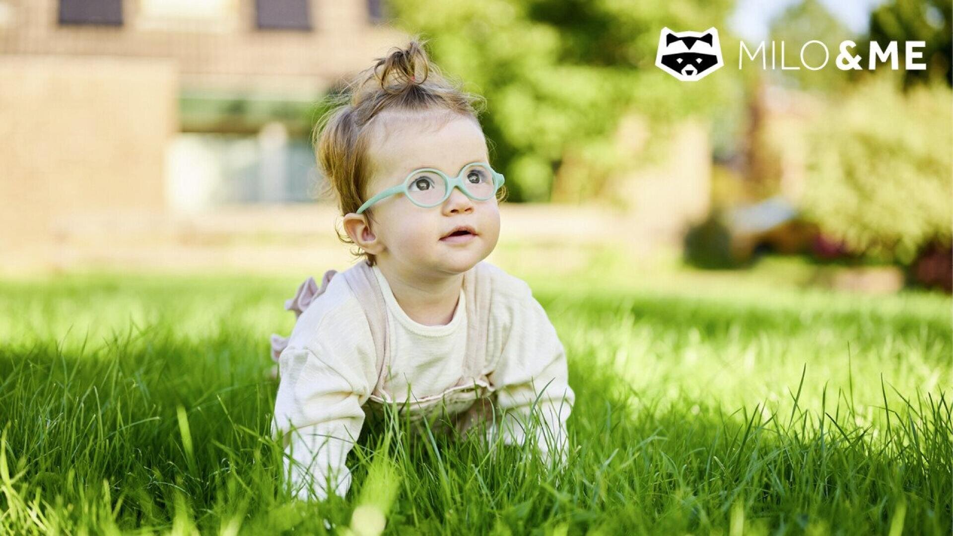 A baby with light-colored hair and green glasses crawls on grass, looking up. The background is blurry, and the logo MILO & ME with a raccoon face is in the top right corner.