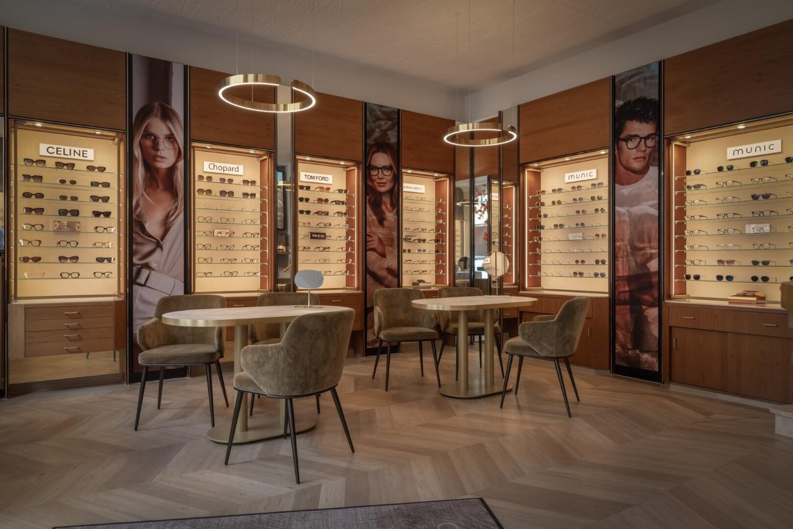 Ein modernes Brillengeschäft mit hellen Holzböden, runden Tischen, beigen Stühlen und beleuchteten Wanddisplays, auf denen verschiedene Brillengestelle und große Modeposter präsentiert werden.