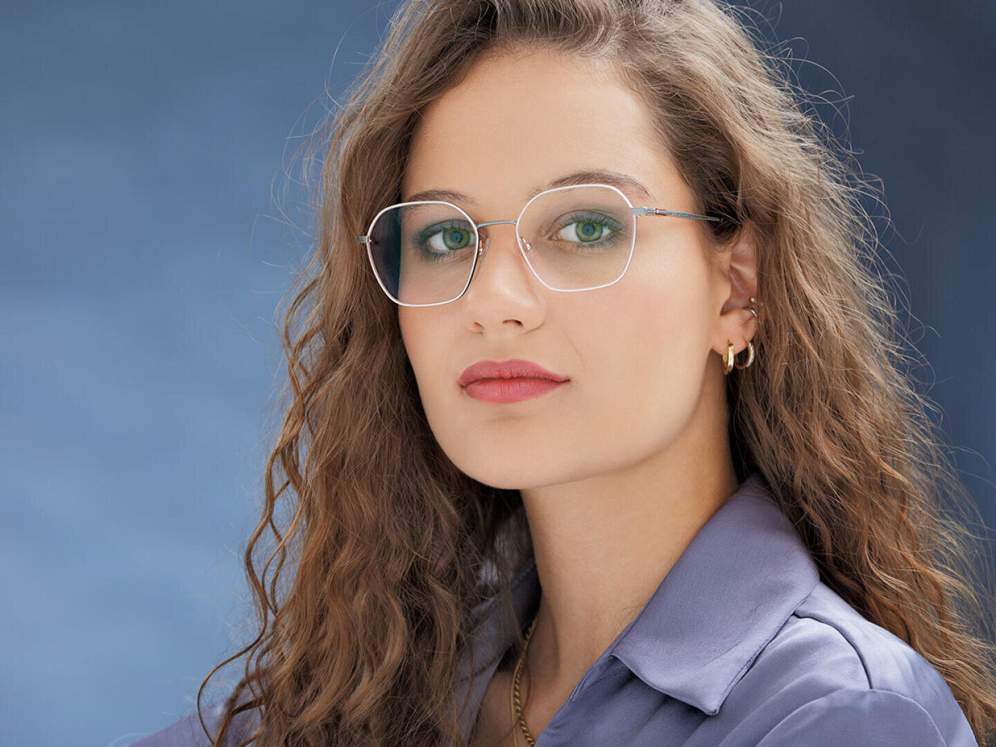 A young woman with long, wavy brown hair wears glasses and gold earrings. She has green eyes, wears light make-up, a lavender collared shirt and poses in front of a blue background.