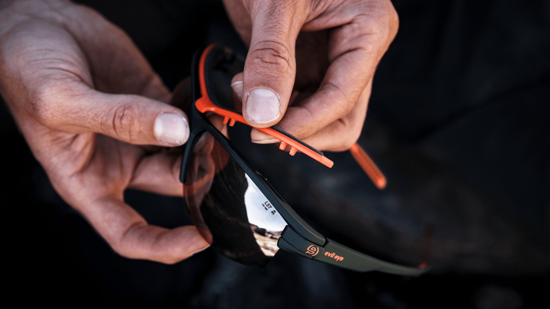 Nahaufnahme von Händen, die eine schwarz-orangefarbene Sport-Sonnenbrille anpassen oder zusammensetzen, wobei eine Hand den Rahmen hält und die andere ein kleines orangefarbenes Teil über dem Glas anbringt.