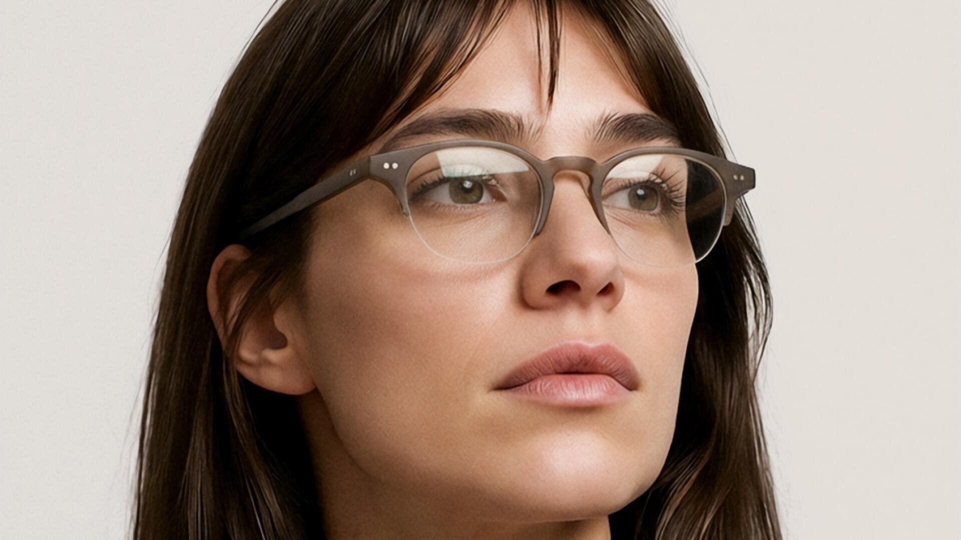 Eine Frau mit langen braunen Haaren, die eine Brille mit klarem Gestell, einen schwarzen Blazer und eine weiße Bluse trägt, schaut vor einem schlichten hellen Hintergrund leicht zur Seite.