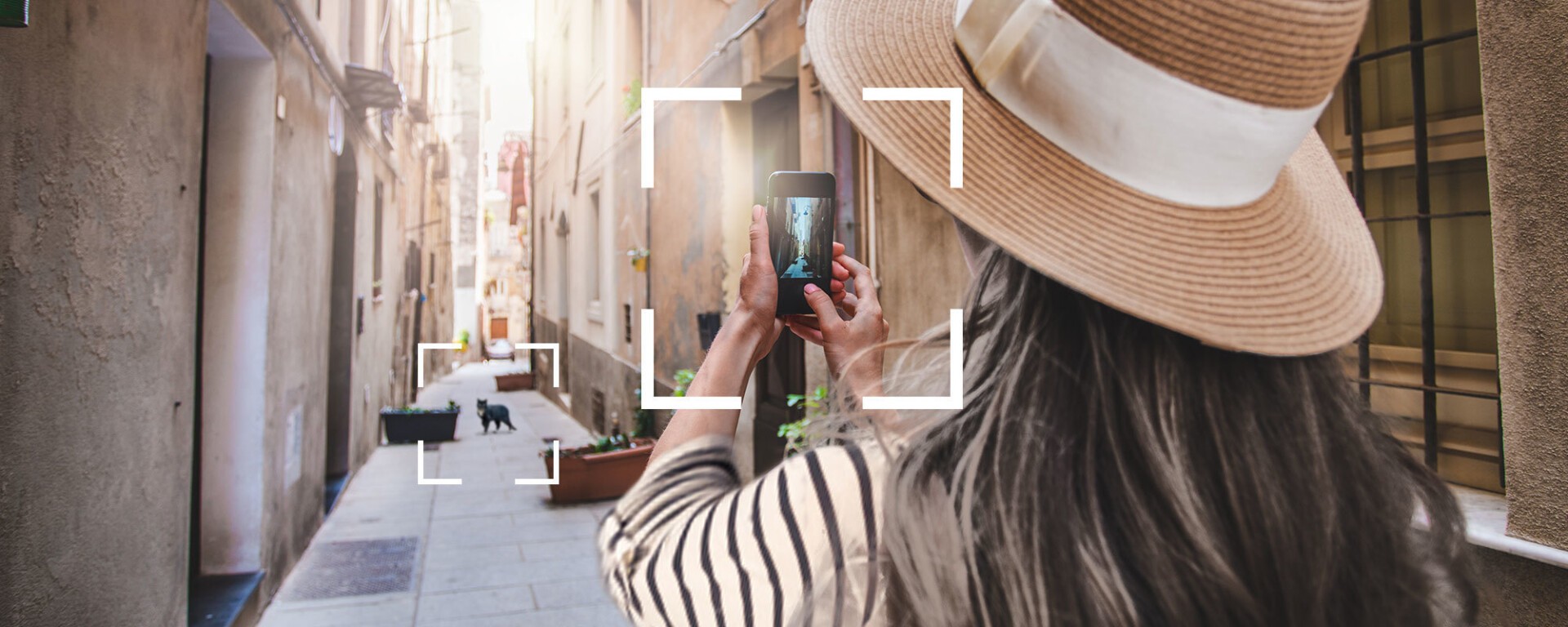 Eine Frau mit breitkrempigem Hut fotografiert in einer schmalen, sonnenbeschienenen Gasse mit ihrem Smartphone. Sie ist von hinten zu sehen, weiter unten in der Straße ist eine kleine schwarze Katze zu sehen. Ihr Handy ist durch eingerahmte Linien hervorgehoben.