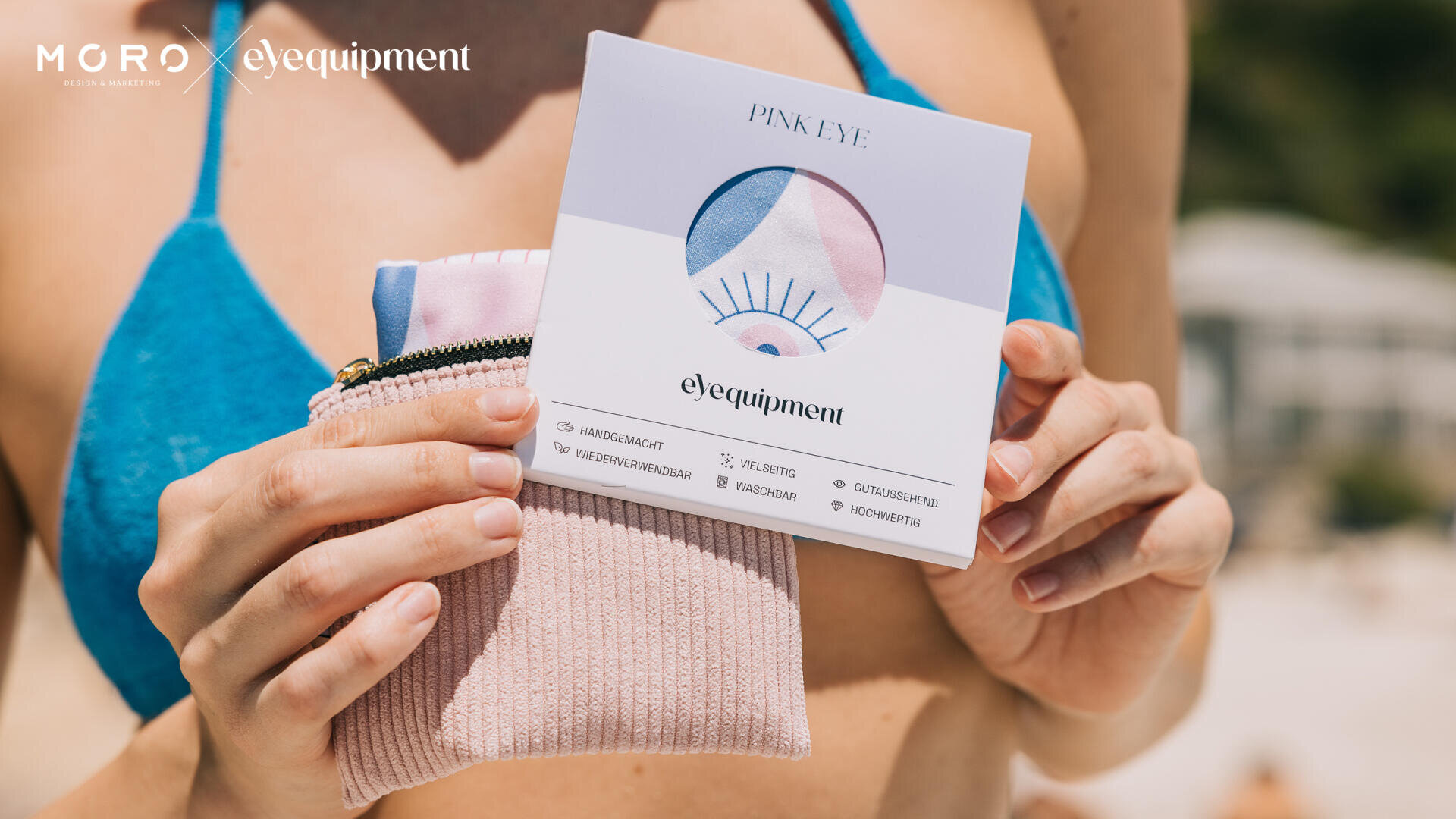 Eine Frau in einem blauen Bikini hält einen rosa Strickbeutel und eine Augenmaske in einer Schachtel mit der Aufschrift PINK EYE von eYequipment by Moro vor einem unscharfen Strandhintergrund.