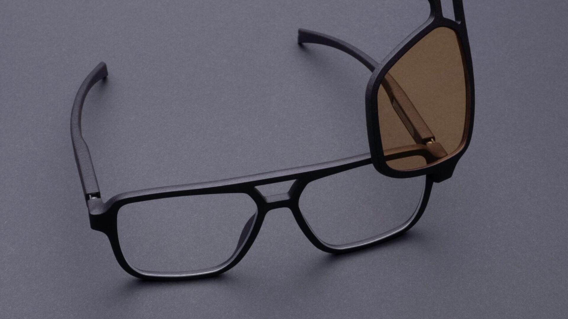 A pair of black eyeglasses with clear lenses lies on a dark surface, overlapped by another pair featuring brown-tinted lenses.