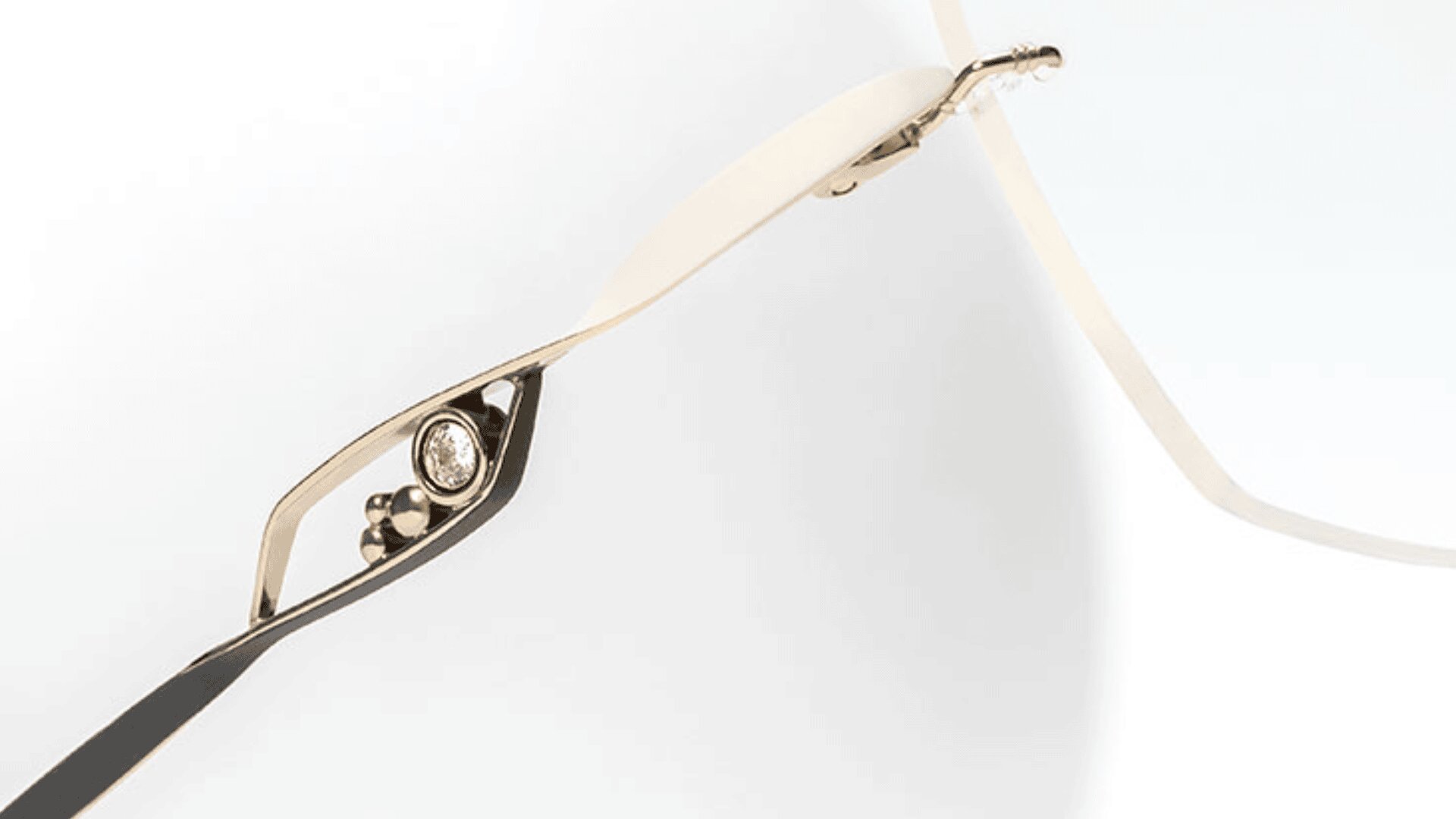 Nahaufnahme der Seite einer Brille mit einem dünnen, goldenen Rahmen und einem kleinen, runden, in den Bügel eingebetteten Diamanten. Das Design ist minimalistisch und modern.
