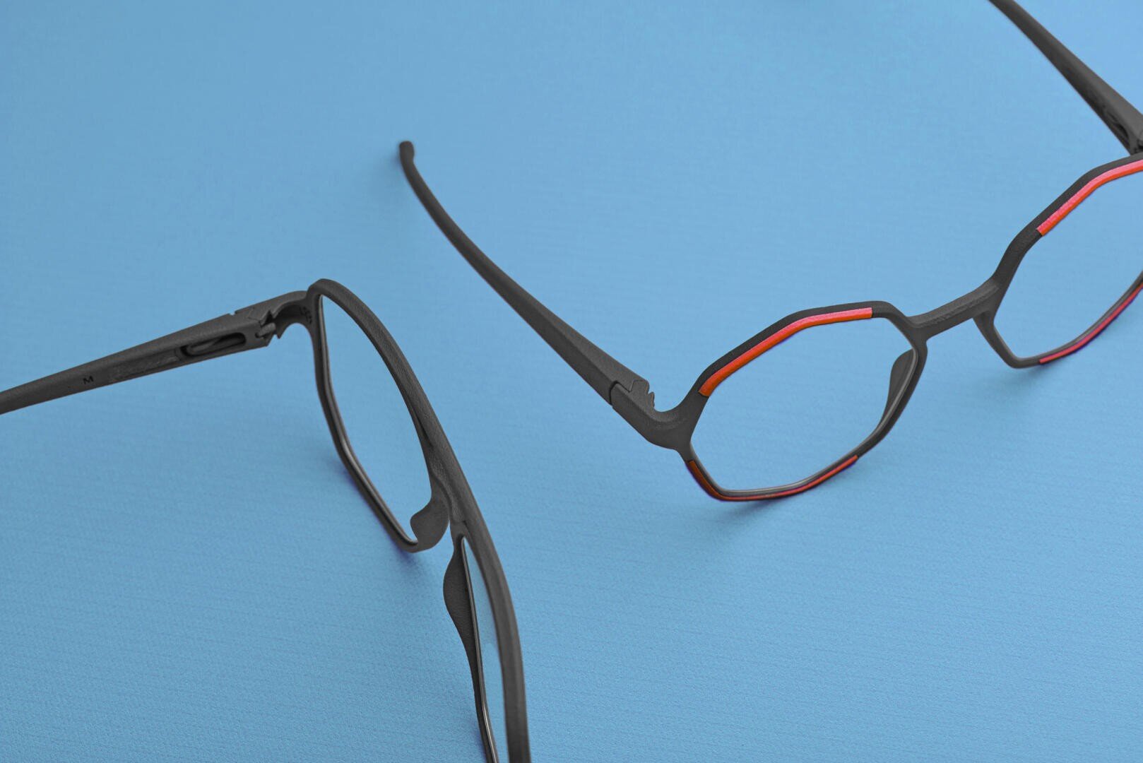 Two pairs of black eyeglasses with geometric frames and red accents are placed on a flat, light blue surface. One pair is fully visible, while the other is partially shown from the side.