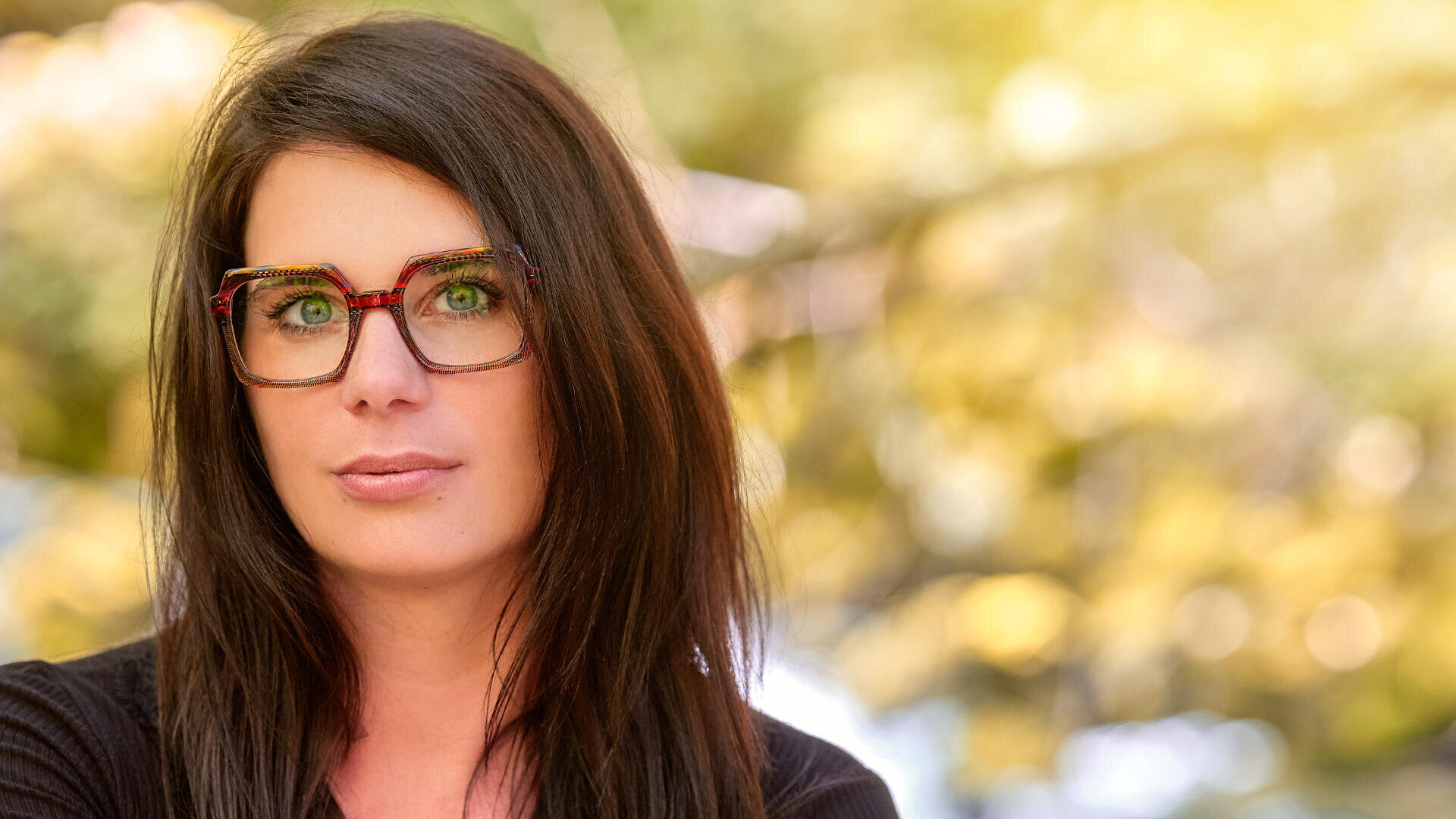 Eine Frau mit langem, dunklem Haar und einer großen, eckigen Brille blickt direkt in die Kamera. Sie befindet sich im Freien vor einem unscharfen, sonnenbeschienenen Hintergrund aus grünem und gelbem Laub.