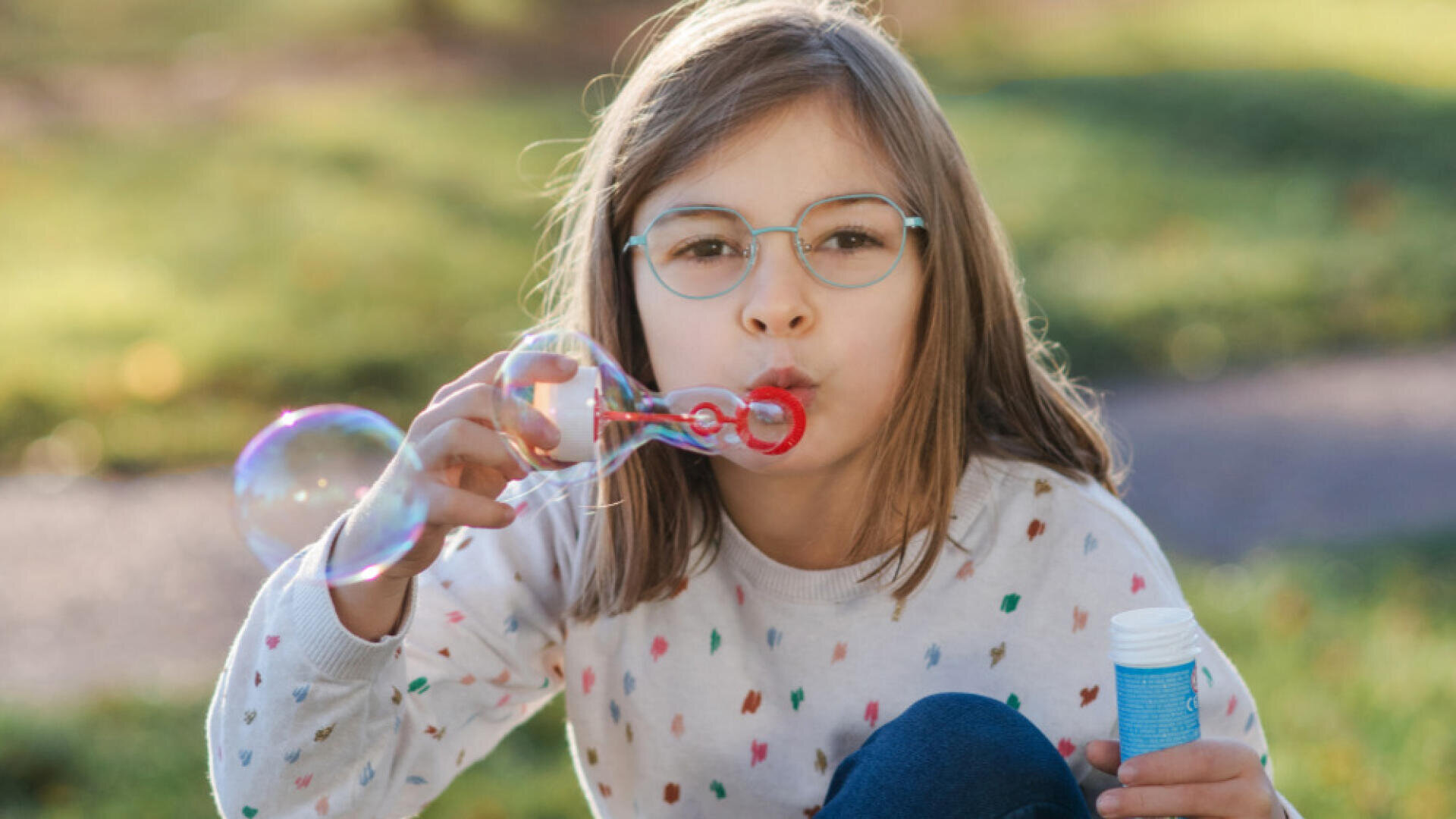 Ein junges Mädchen mit Brille und gemustertem Pullover pustet im Freien Seifenblasen, in der Hand einen Seifenblasenstab und eine blaue Flasche, mit grünem Gras im Hintergrund.