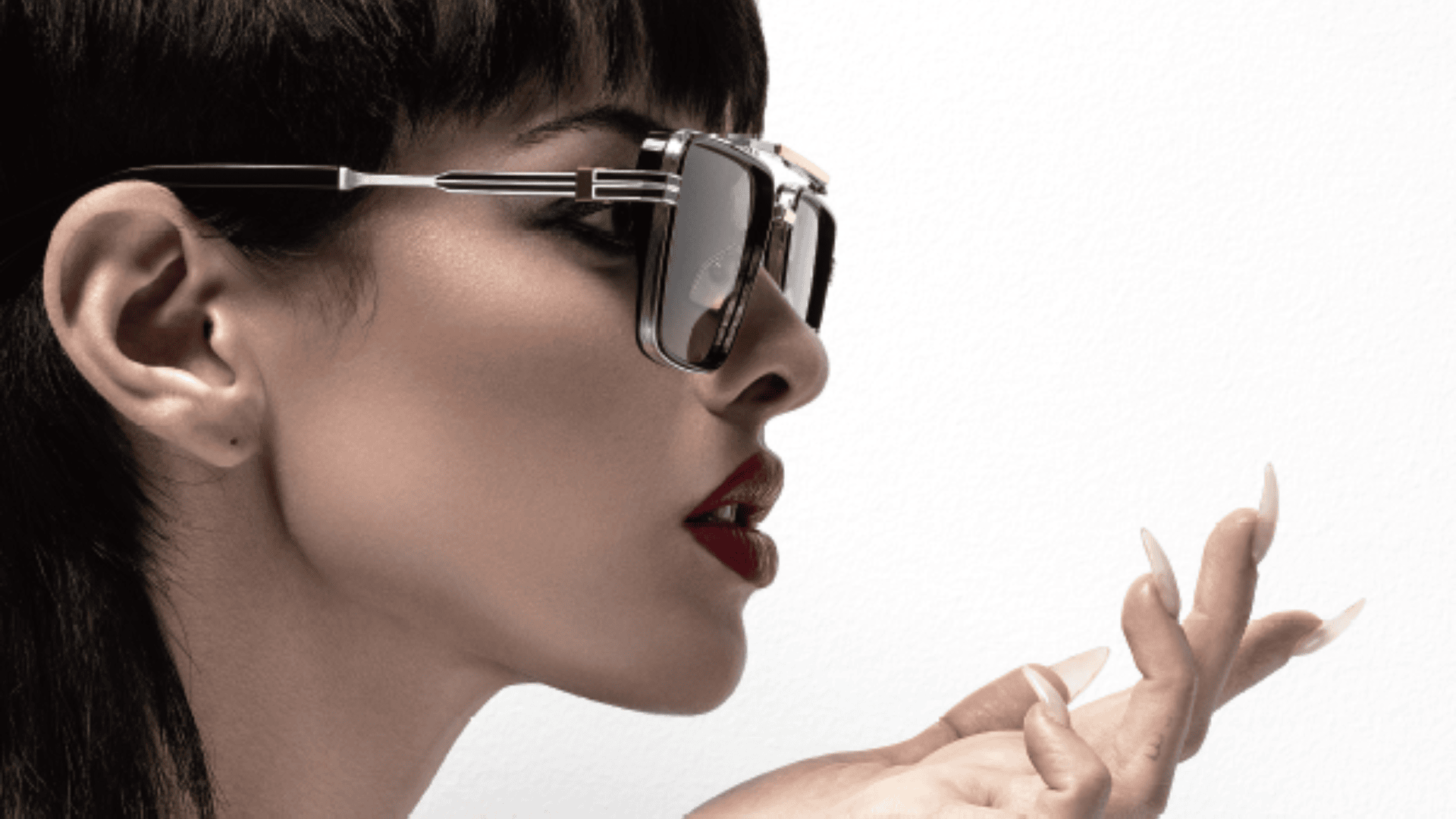 Eine Frau mit dunklem Haar und rotem Lippenstift trägt eine große, stylische Sonnenbrille und lange, manikürte Nägel und hält ihre Hand vor einem weißen Hintergrund in die Nähe ihrer Lippen.