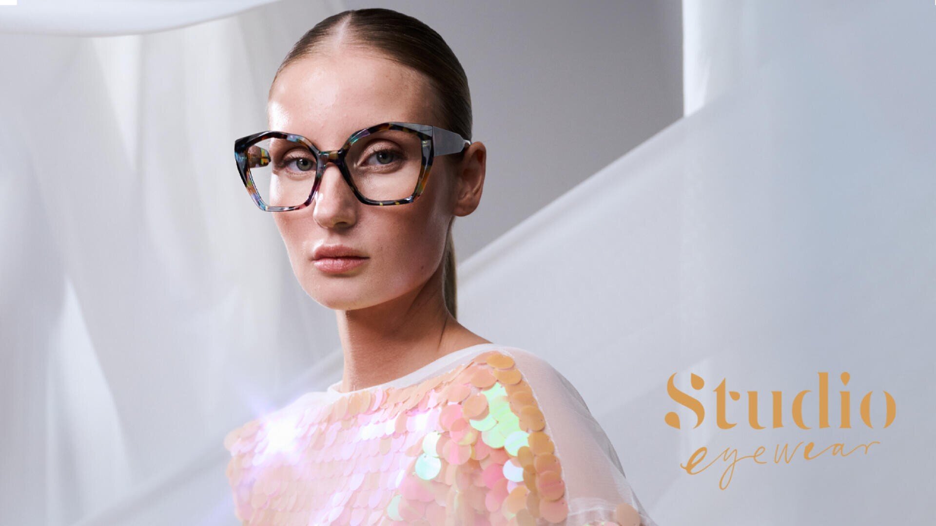Eine Frau mit glattem Haar trägt eine große, modische Brille in Schildpattoptik und ein Oberteil mit schillernden Pailletten und posiert in einer sanft beleuchteten, modernen Umgebung. Auf dem Bild steht in goldener Schrift „Studio Eyewear“.
