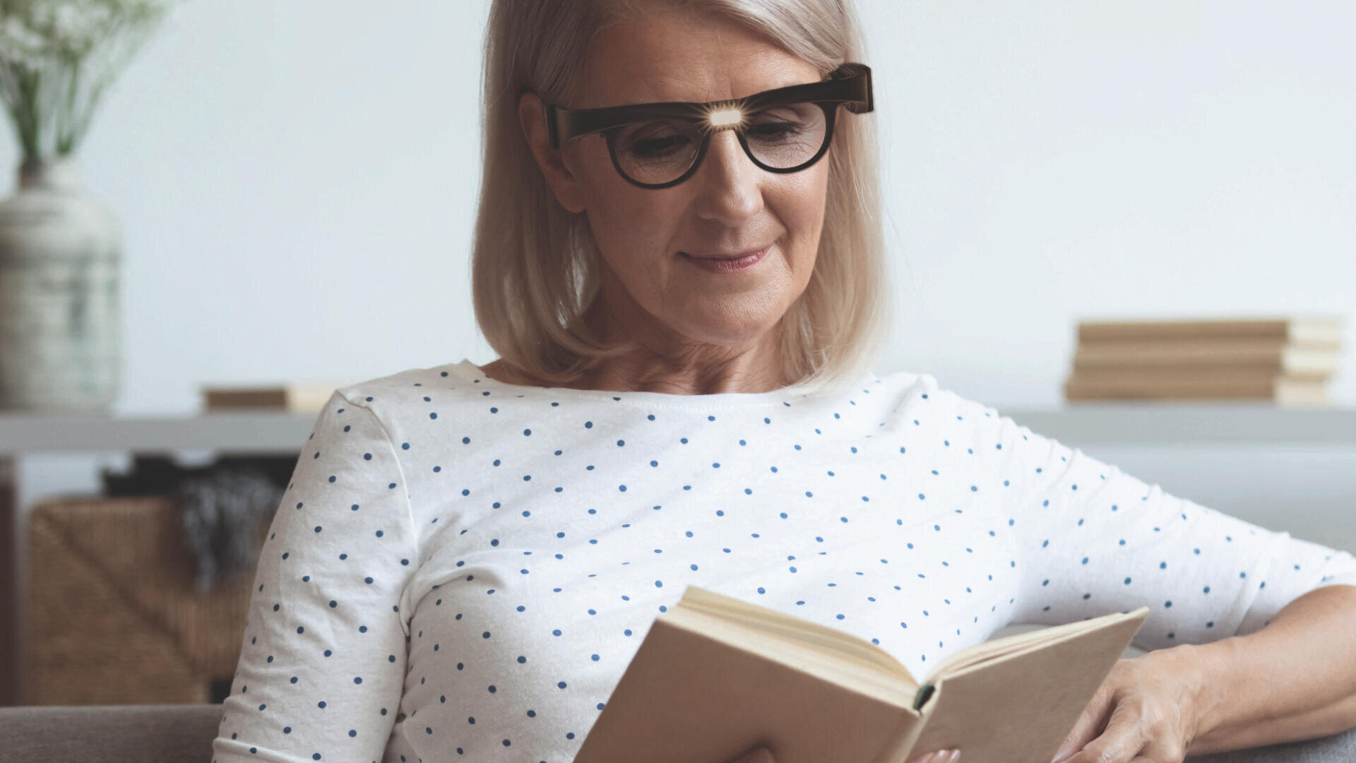 Eine Frau mit blonden Haaren und Brille, die ein weißes Hemd mit schwarzen Punkten trägt, sitzt auf einem Sofa und liest ein Buch. Im Hintergrund stehen eine Vase mit Blumen und ein Bücherstapel auf einem Regal.