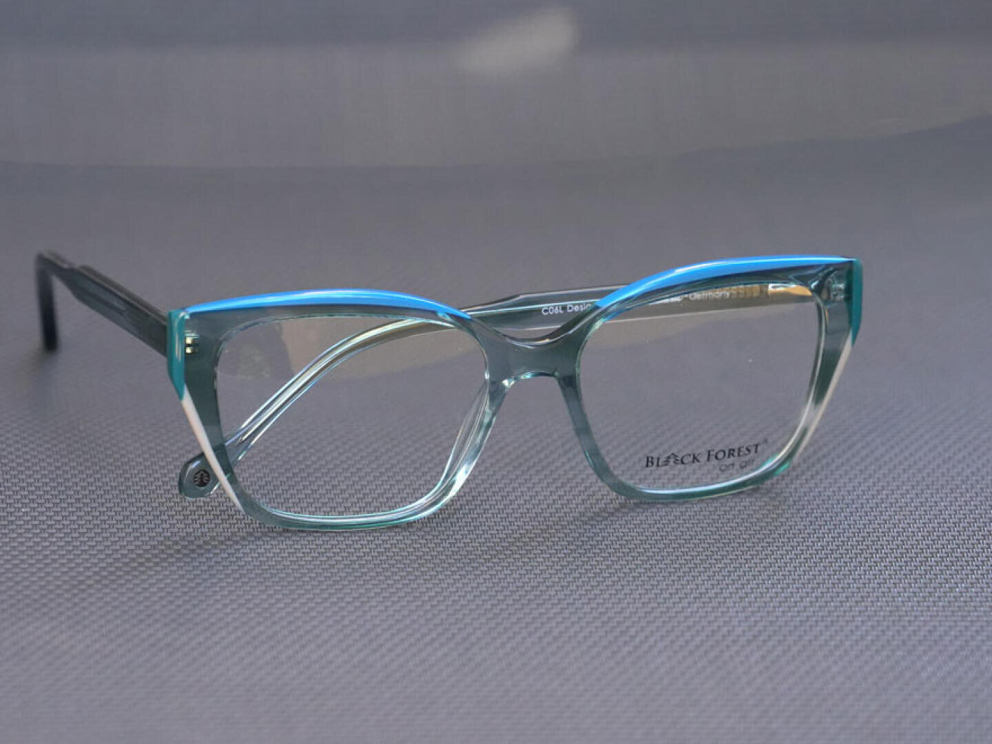 Eine hellblaue Katzenaugenbrille mit klaren Gläsern ruht auf einer strukturierten grauen Oberfläche. Die Innenseite eines Glases ist mit dem Markennamen BLACK FOREST gekennzeichnet.