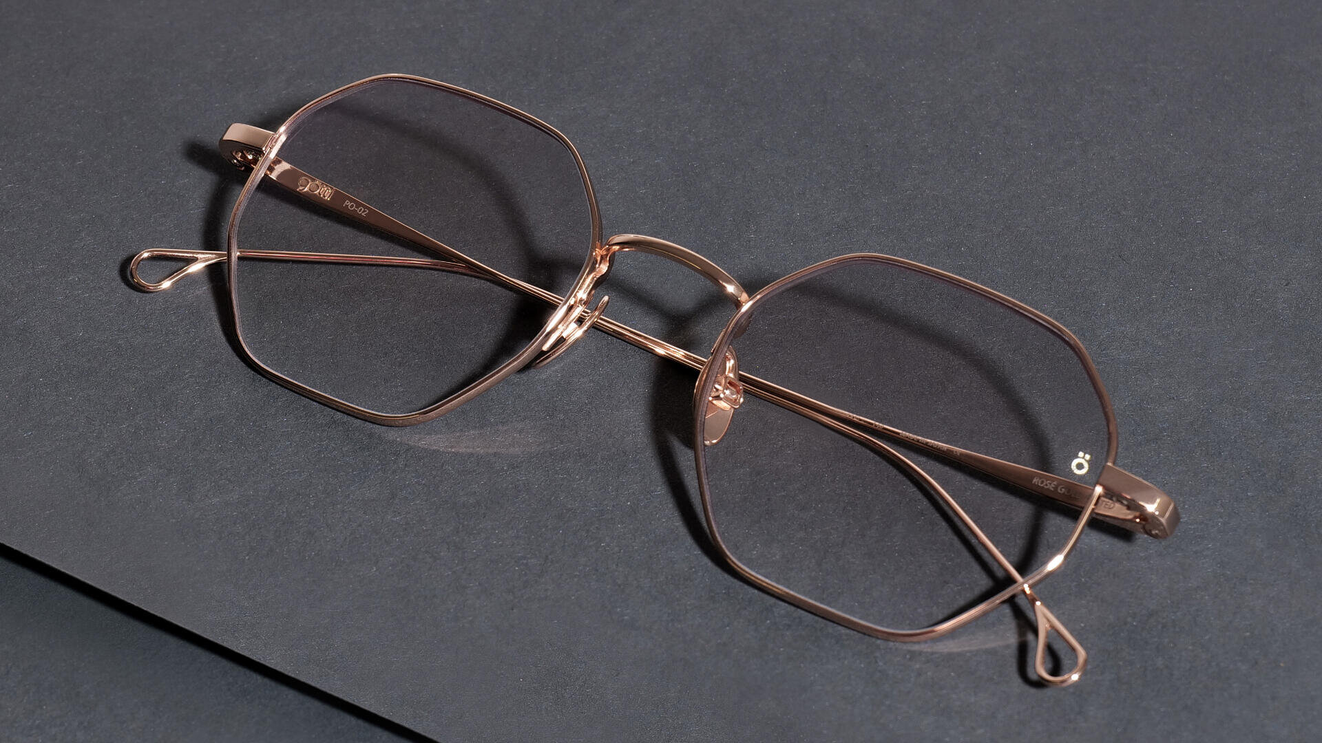 Eine goldene Brille mit geometrischer Form und dünnem Metallrahmen liegt auf einer dunklen, strukturierten Oberfläche.