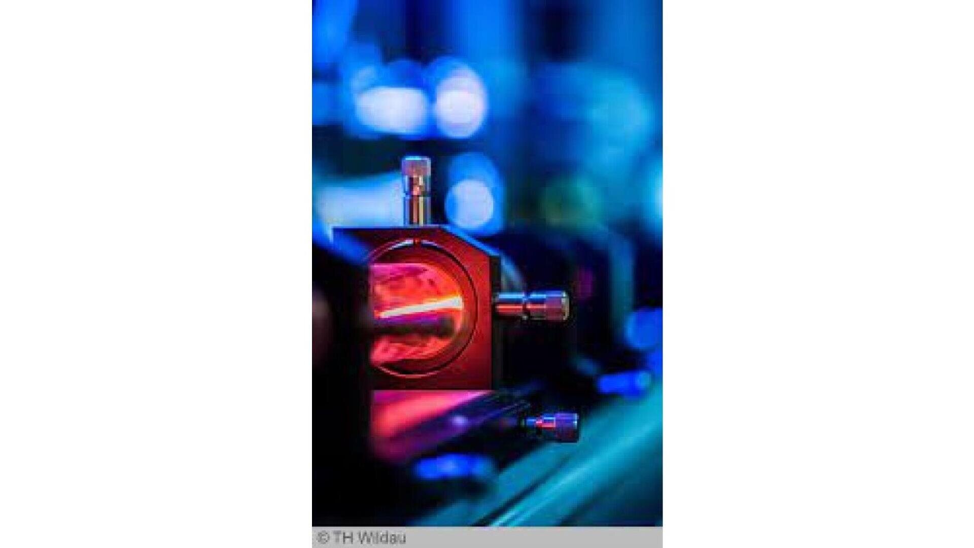 Close-up of scientific equipment with a glowing red light inside, surrounded by bolts and metallic parts, set against a blurred blue and purple background.