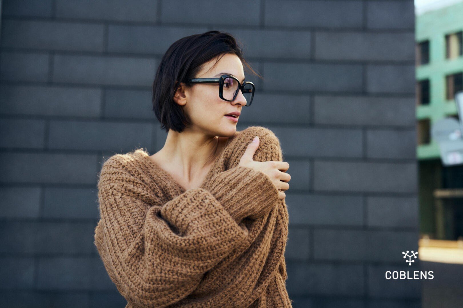 Eine Person mit kurzen dunklen Haaren und einer großen schwarzen Brille trägt einen dicken braunen Pullover und steht vor einer dunklen Backsteinwand. Die Person blickt zur Seite und hält den Pullover fest. In der Ecke erscheint das COBLENS-Logo.