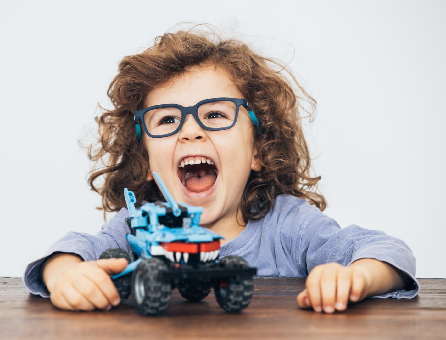 Ein kleines Kind mit lockigem Haar und Brille sitzt fröhlich lachend an einem Tisch und spielt mit einem blauen Spielzeugauto, auf dessen Vorderseite scharfe Zähne aufgemalt sind.