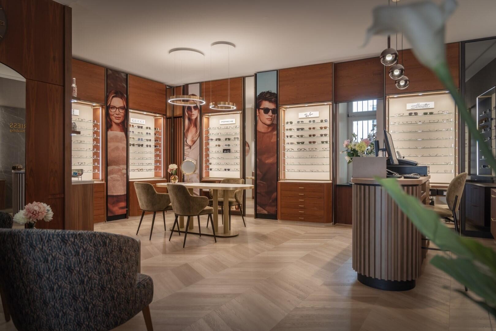 Ein modernes Optikergeschäft mit Holzböden, stilvollen Stühlen, Brillenvitrinen, großen Modepostern und Pendelleuchten, die eine warme, einladende Atmosphäre schaffen.
