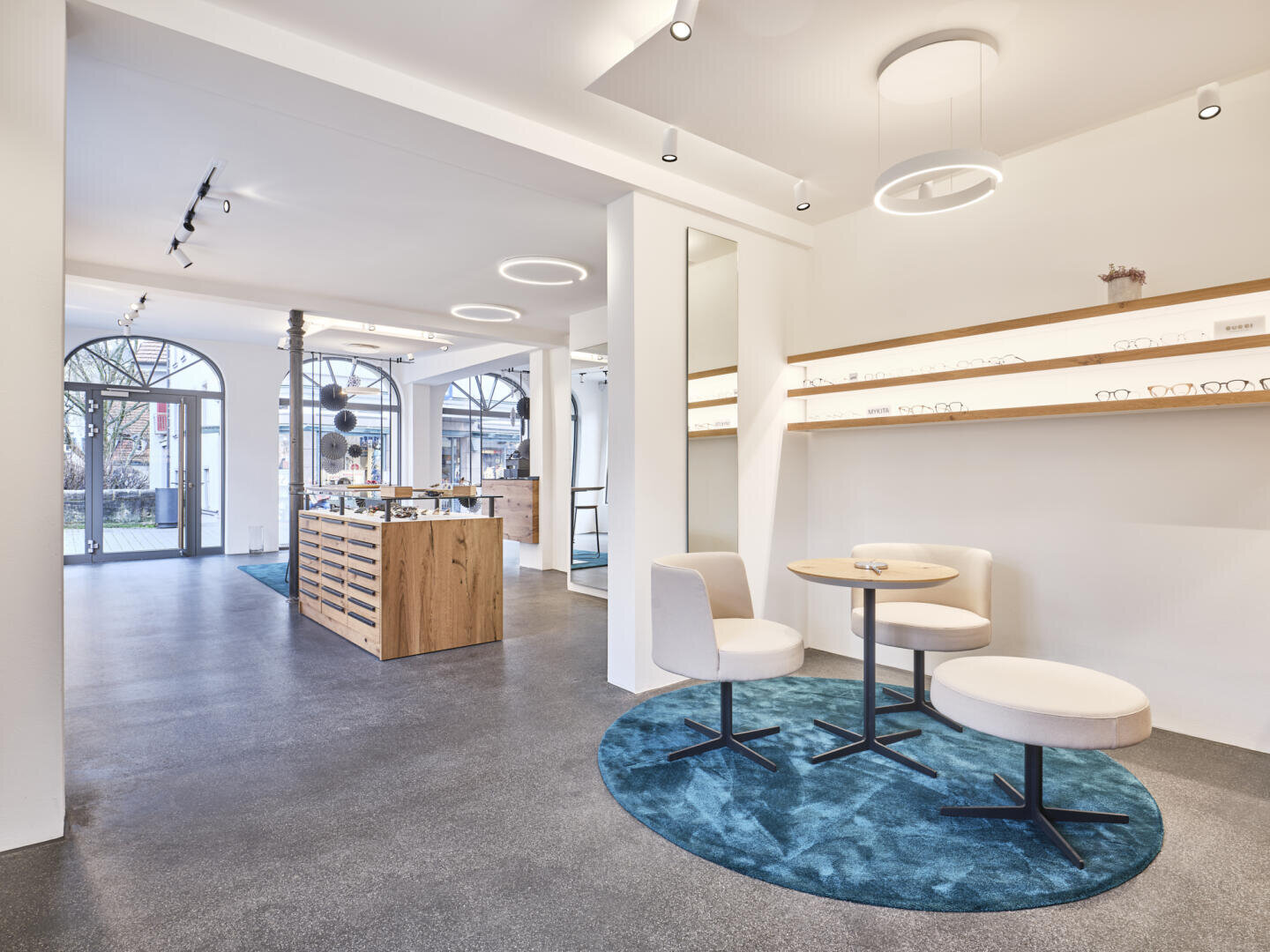 Ein modernes Optikergeschäft mit weißen Wänden, runden Deckenleuchten und großen Bogenfenstern. Es gibt Ausstellungsregale mit Brillen, Holztheken und zwei runde Tische mit weißen Stühlen auf blauen Teppichen.