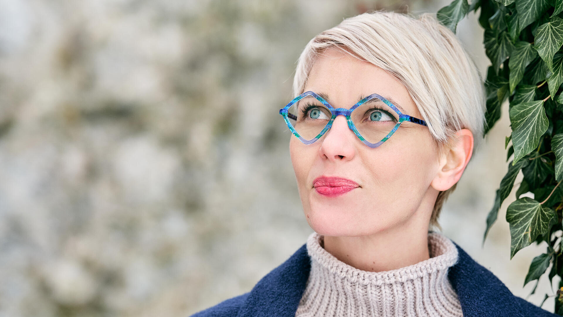 Eine Frau mit kurzen blonden Haaren und einer blauen Brille mit geometrischem Muster blickt nachdenklich auf. Sie trägt einen cremefarbenen Rollkragenpullover und einen blauen Mantel und steht im Freien neben grünen Efeublättern.