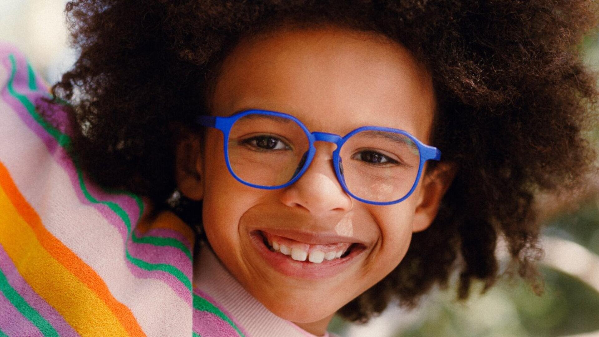 A smiling child with curly hair wears bright blue glasses and a colorful striped sweater. The background is softly blurred, highlighting the childs joyful expression.
