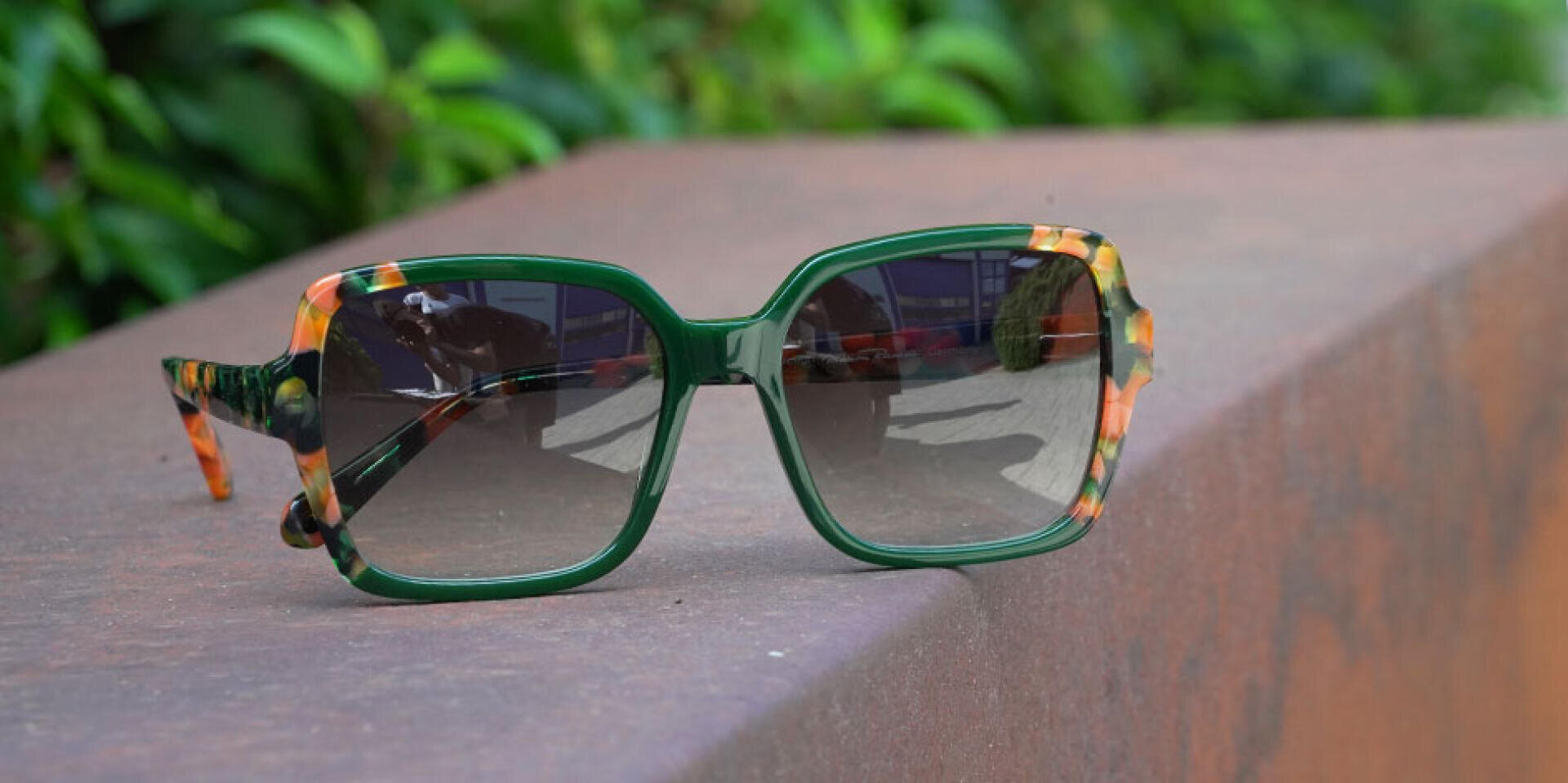 A pair of oversized sunglasses with green and orange marbled frames rests on a brown ledge, with green foliage blurred in the background.