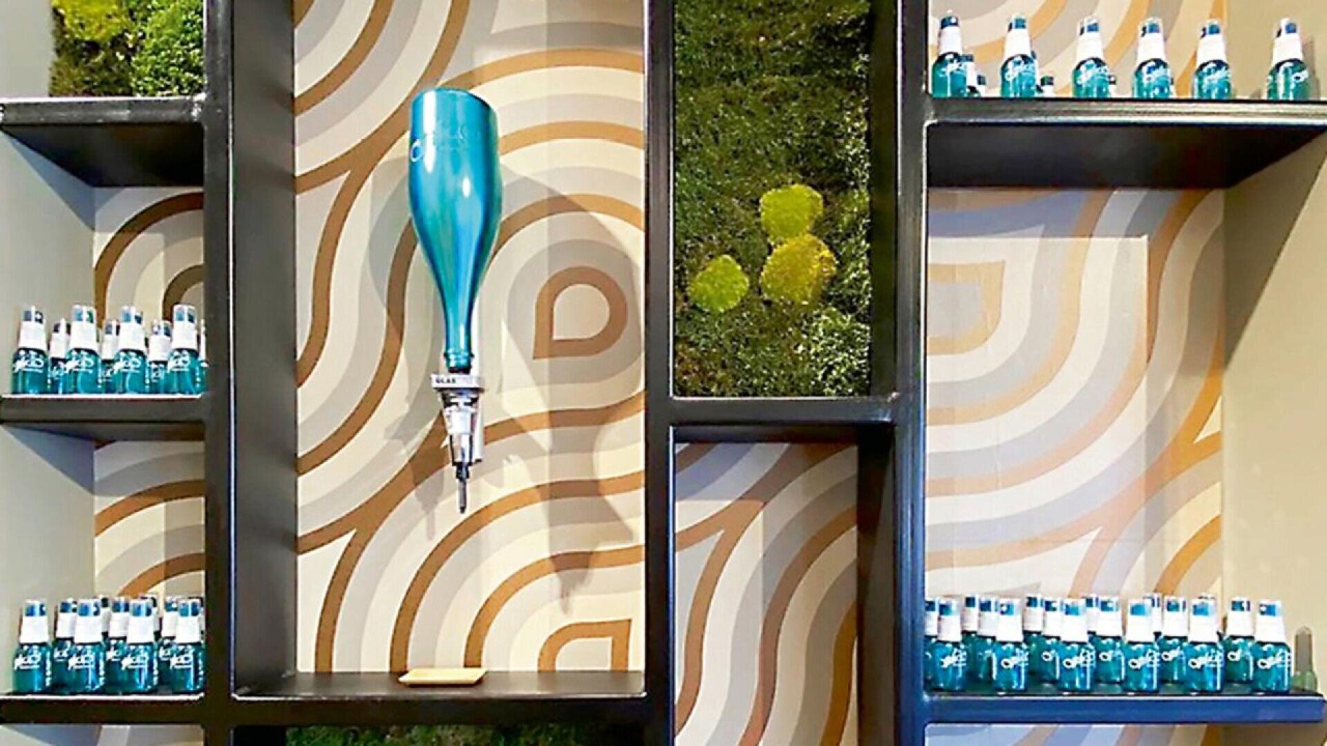 Wall shelves display small blue bottles, a blue beverage dispenser, and patches of green moss, set against a retro-patterned wallpaper with brown and beige circular designs.