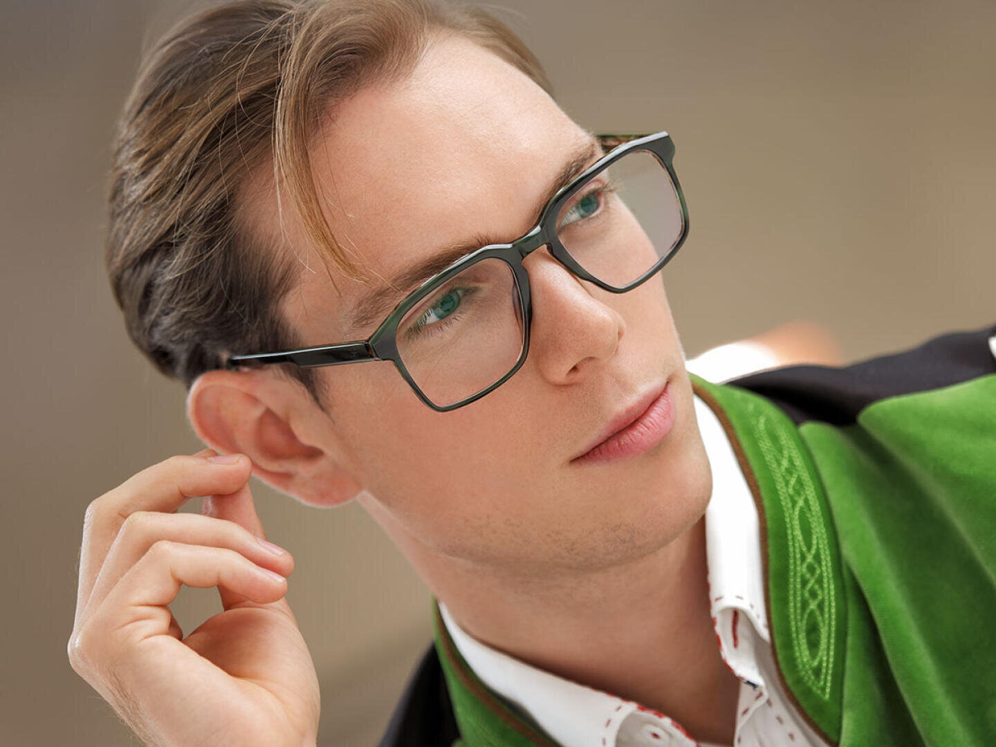 A young man with light brown hair and glasses looks to the side. He is wearing a green jacket over a white collared shirt with red accents. He has a thoughtful expression on his face and is touching his ear. The background is blurred.