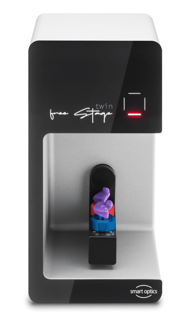 A compact, modern benchtop device called free Stage twin from Smart Optics, featuring a small platform with a blue and red sample holder and a purple tooth shape inside the device.