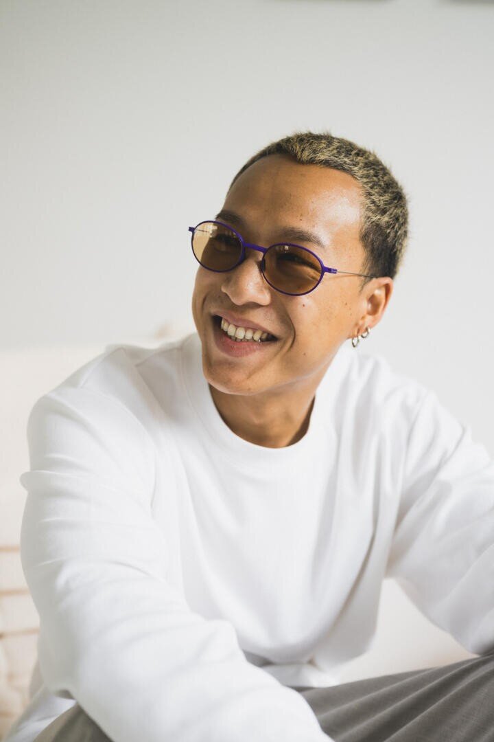 Eine lächelnde Person, die eine runde lila Brille, goldene Ohrringe und ein weißes langärmeliges Hemd trägt, sitzt in einem Haus vor einem hellen Hintergrund.