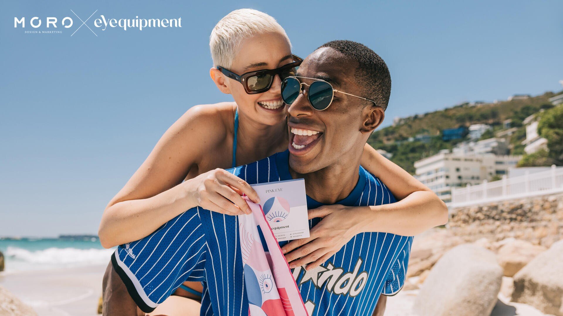 Eine lächelnde Frau mit kurzen blonden Haaren umarmt einen lachenden Mann von hinten an einem sonnigen Strand. Beide tragen Sonnenbrillen und blau gestreifte Hemden. Der Mann hält eine rosa-weiße Produktverpackung in der Hand. Im Hintergrund sind Gebäude und Hügel zu sehen.