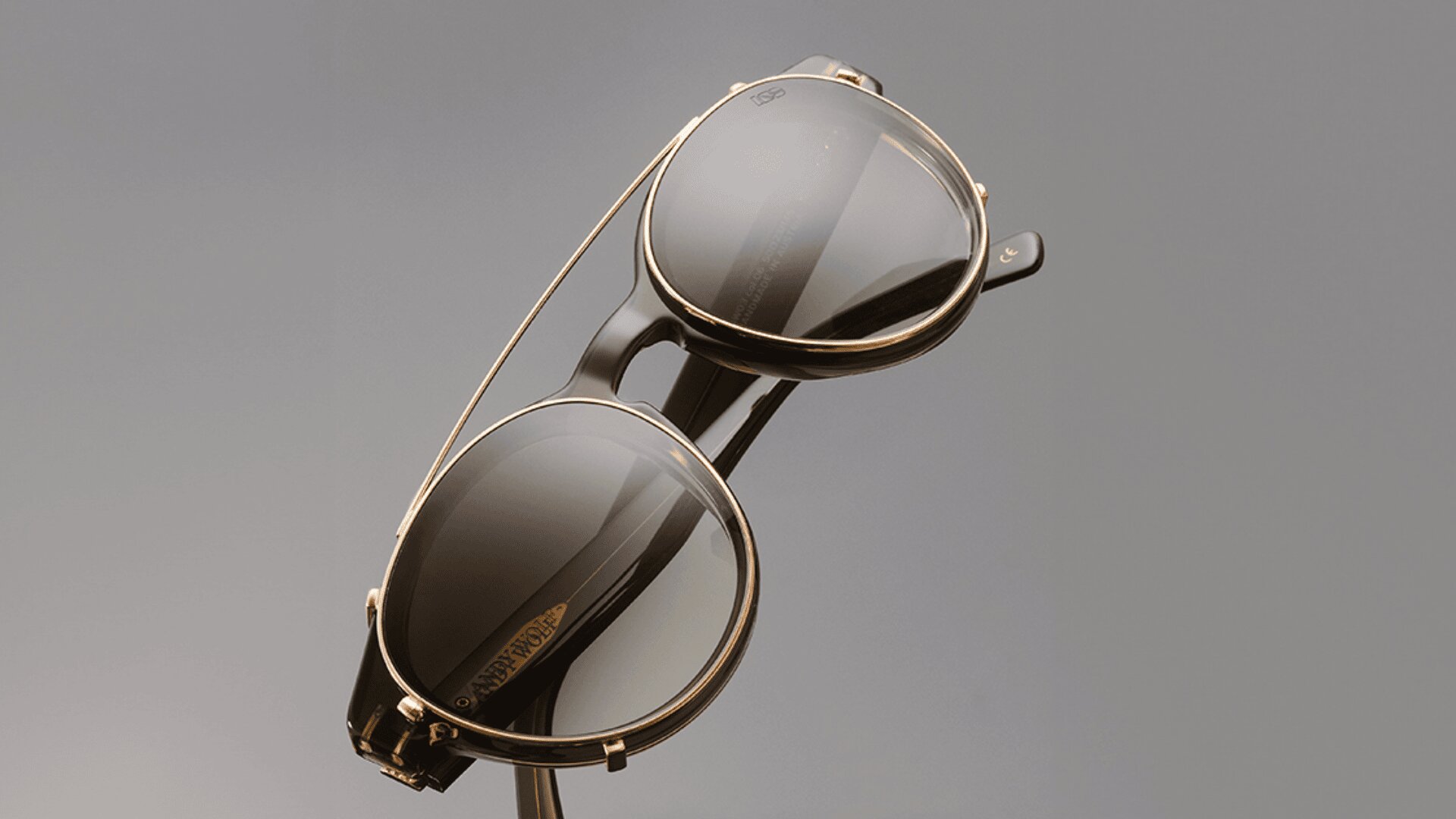 Eine stilvolle Sonnenbrille mit runden, dunklen Gläsern und einem dünnen, goldenen Rahmen, der sich schräg vor einem schlichten grauen Hintergrund abhebt.