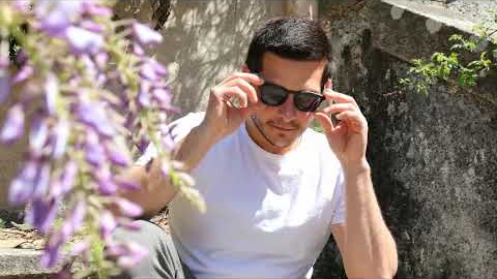 A man wearing a white t-shirt sits outdoors on stone steps, adjusting his black sunglasses. Purple flowers are visible in the foreground, slightly out of focus. Sunlight casts shadows on the scene.