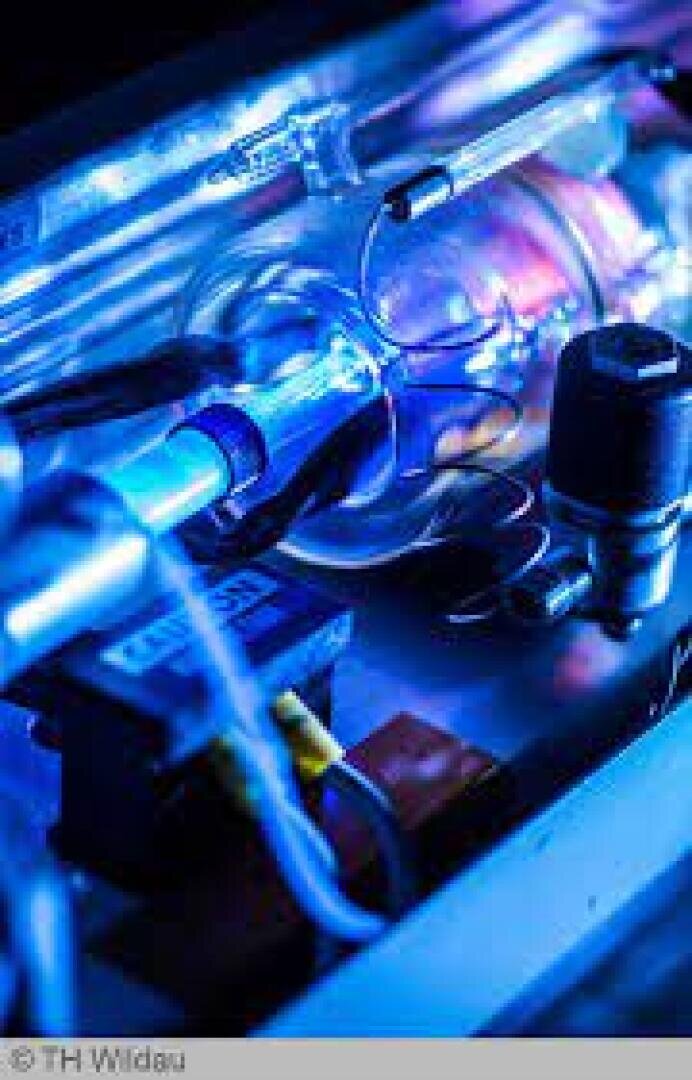 Nahaufnahme einer wissenschaftlichen Laborausrüstung, einschließlich Glasröhren, metallischer Komponenten und elektronischer Teile, beleuchtet von blauem und violettem Licht.