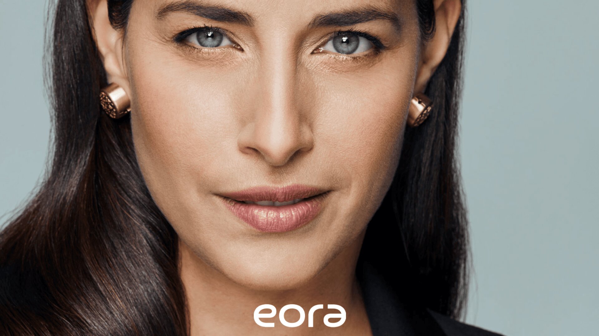 Nahaufnahme einer Frau mit langem dunklem Haar, hellen Augen und dezentem Make-up, die goldene Ohrringe und ein schwarzes Oberteil trägt, darunter steht in weißer Schrift das Wort „Eora“.