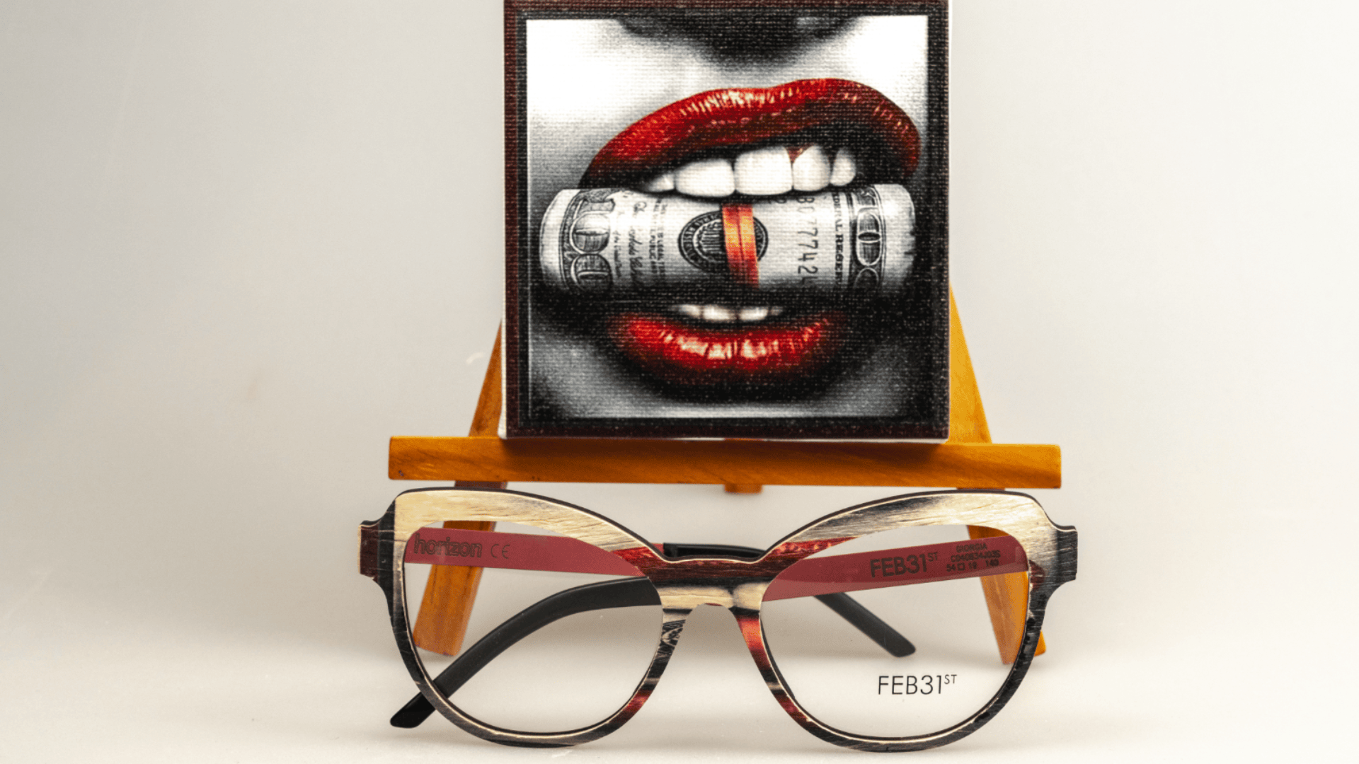 Eine Brille steht vor einer kleinen Staffelei, auf der rote Lippen zu sehen sind, die in eine aufgerollte Dollarnote beißen, alles vor einem schlichten weißen Hintergrund.