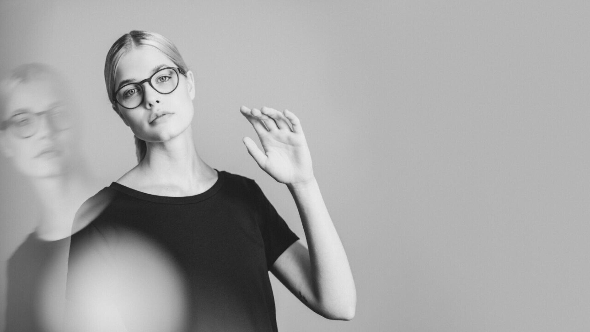 Black and white photo of a woman with light hair in a ponytail, wearing glasses and a dark shirt, raising one hand. Her expression is neutral, and there is a blurred reflection of her on the left side of the image.