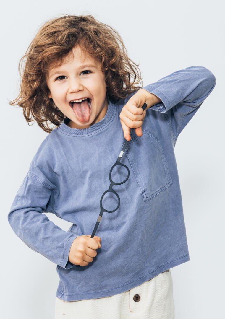 Ein kleines Kind mit lockigem Haar und blauem Hemd streckt spielerisch die Zunge heraus und hält dabei eine runde schwarze Brille am Stiel vor der Brust.