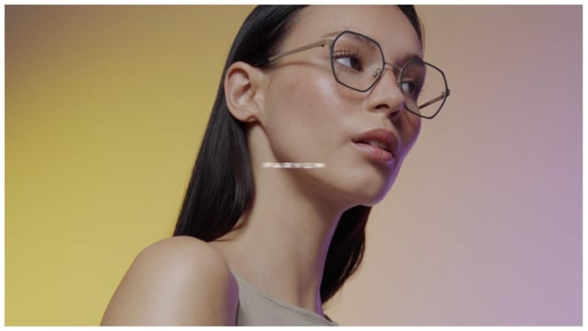 Eine Frau mit langen dunklen Haaren, die eine große runde Brille und ein ärmelloses Oberteil trägt, blickt vor einem gelb-violetten Hintergrund mit Farbverlauf leicht nach oben.